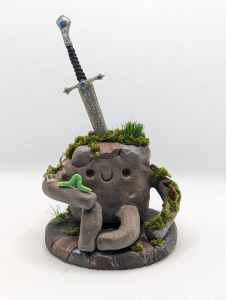 A happy gray little rock guy with a sword protruding from his back. He is covered in moss and crystal chips and has an inch worm on his hand. He is smiling and happy. He sits on a flagstone base with bits of moss. He is a handmade polymer clay figurine. He has a few accents of antique bronze or copper color on his mostly granite gray exterior.